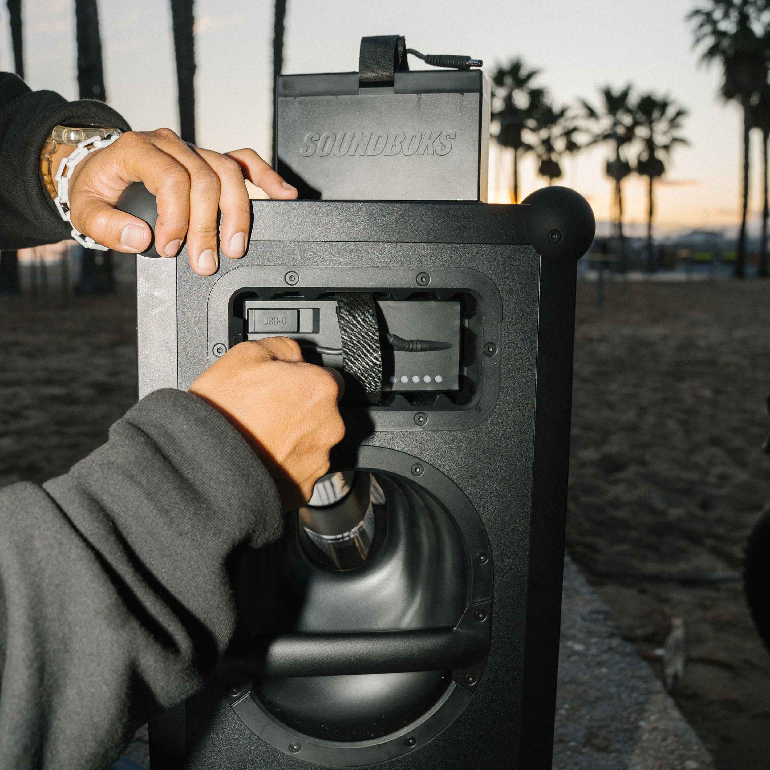 Close-up of a person inserting THE BATTERY into a black SOUNDBOKS 4 party speaker outdoors at sunset. The background features palm trees and a beach setting. The person is wearing a dark sweatshirt and a wristwatch, preparing for an evening of music by the shore